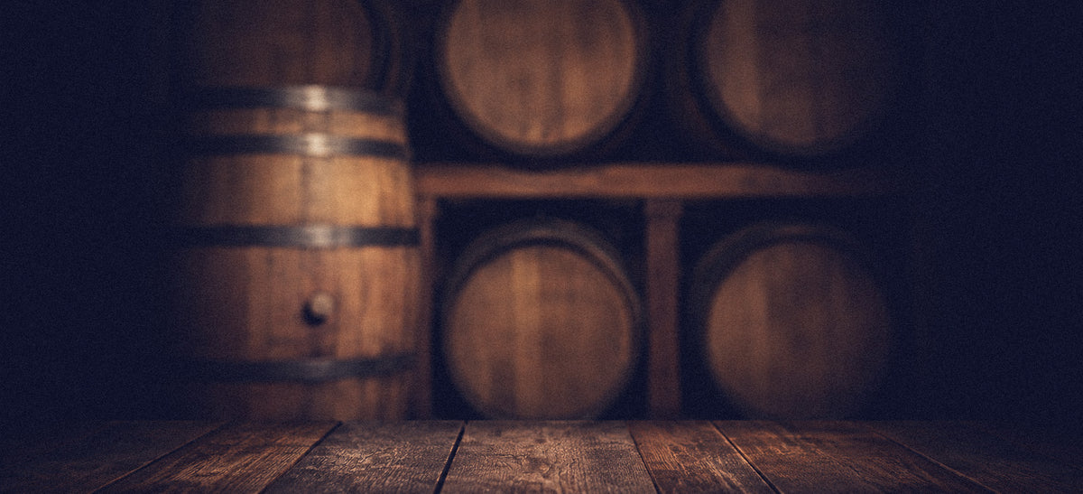 A rustic wooden barrel with metal bands stands on a dark wood floor in a dimly lit cellar, with several more wooden barrels stacked in the background.