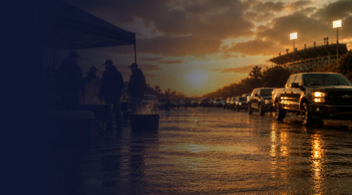 A wet road at sunset with several cars parked along the right side, a dark canopy and silhouetted people on the left, and stadium lights in the distance under a dramatic orange sky.