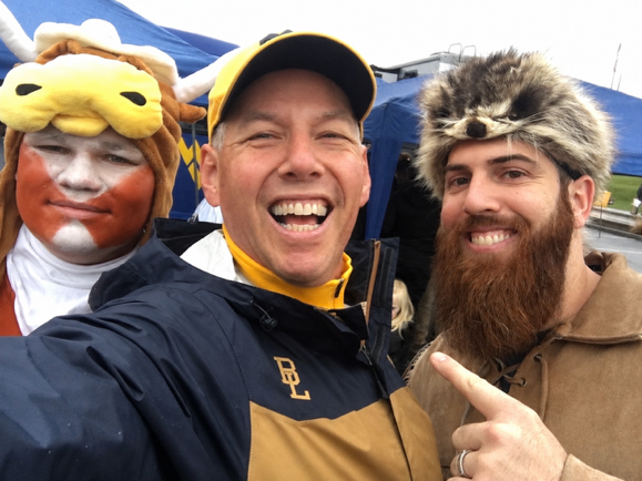 Three men taking a selfie outdoors under a blue tent. The man in the center wears a navy and tan jacket with a gold 'EL' logo and a yellow cap.