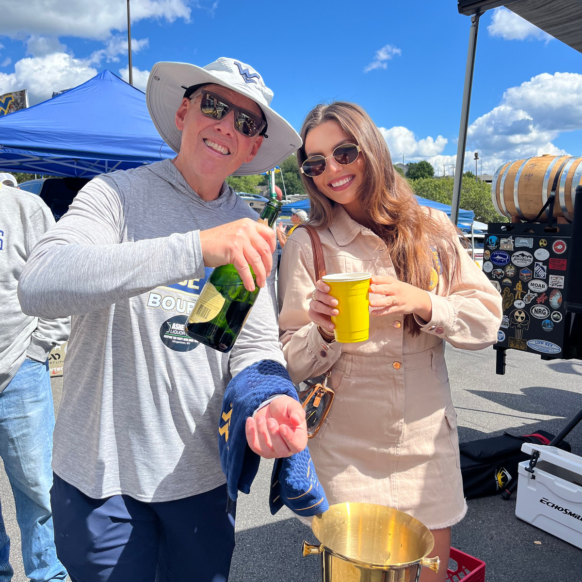 A man in a white hat pouring champagne from a green bottle into a brass bucket, standing next to a woman in a beige dress holding a yellow cup at an outdoor event under a blue sky.